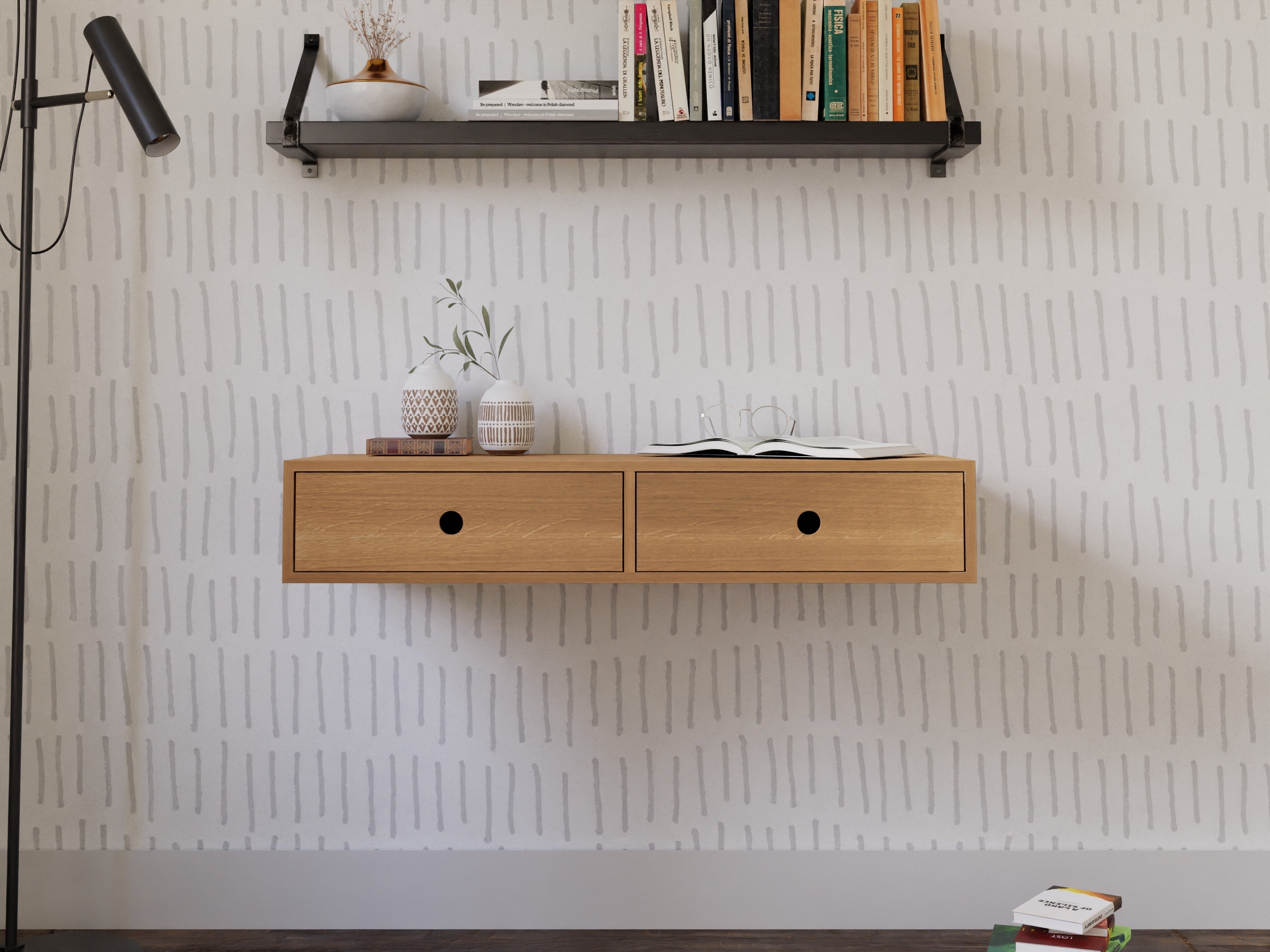 A minimalist floating wooden shelf with two drawers is mounted on a wall with striped wallpaper. An open book and potted plants are on the shelf, with a wall lamp and books on a higher shelf above.
