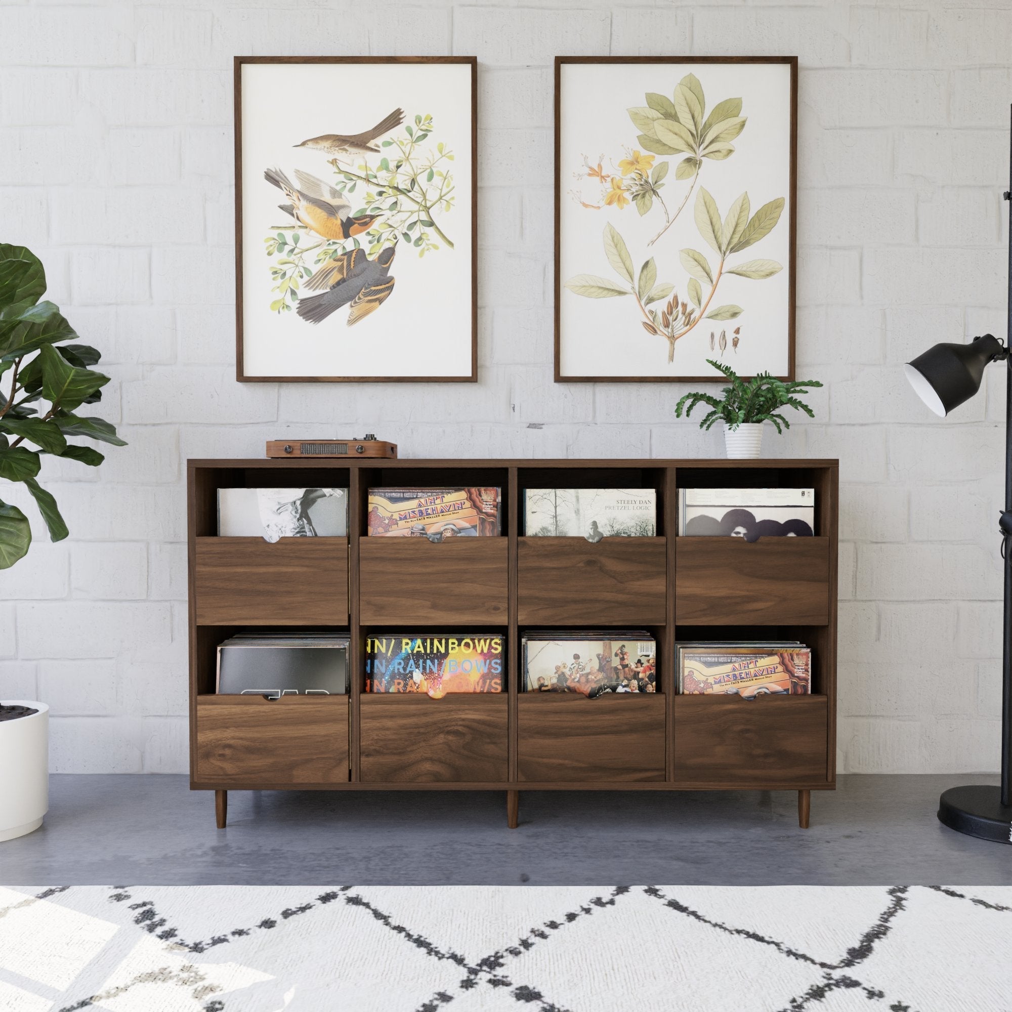A wooden cabinet with multiple shelves features vinyl records and decor items, set against a white brick wall with two botanical framed prints above. A potted plant and floor lamp are on either side.
