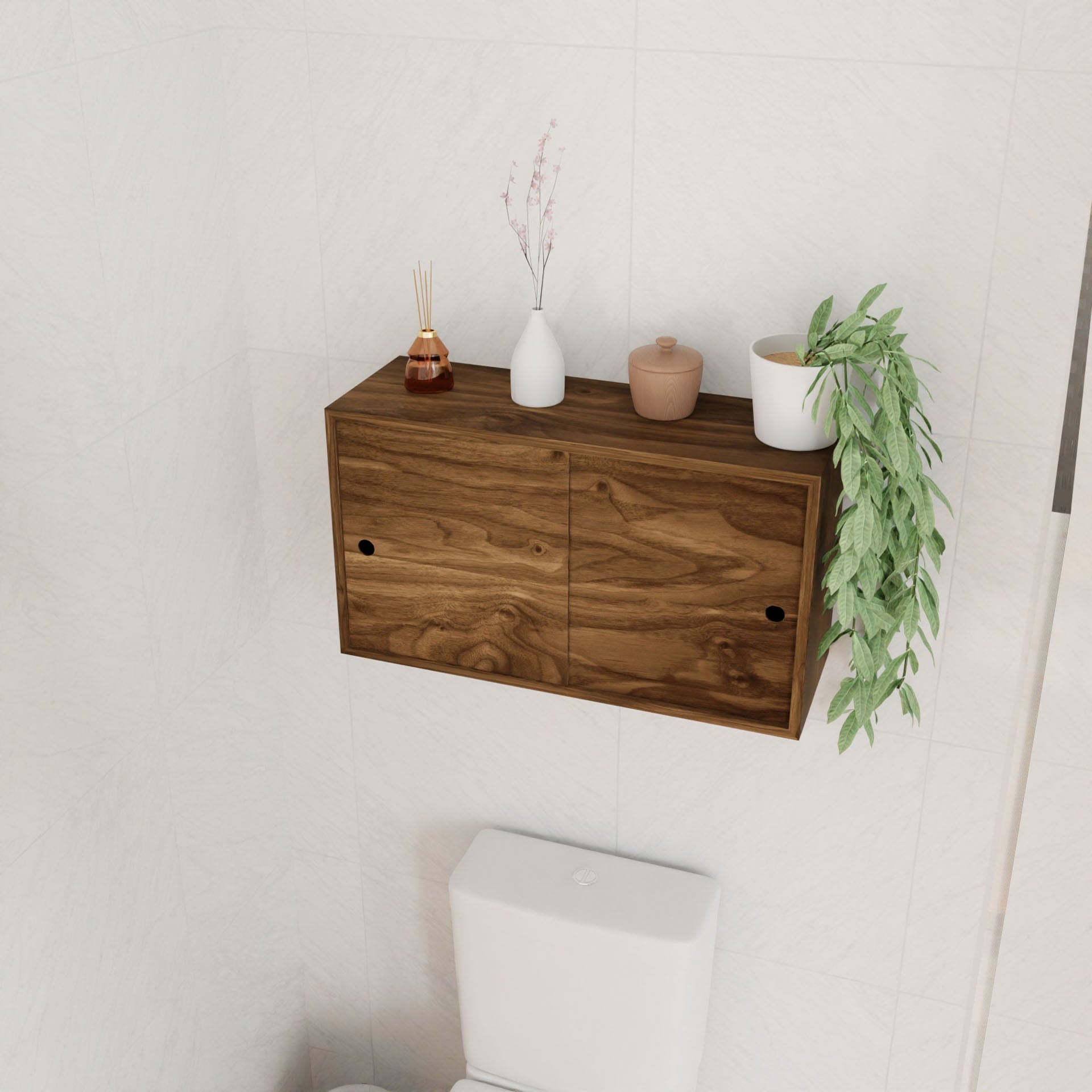 A Krovel Cupboard with Shelf in Walnut is wall mounted above a white toilet. Decorative items like vases, a jar, and a trailing potted plant are displayed on top of the floating cabinet.