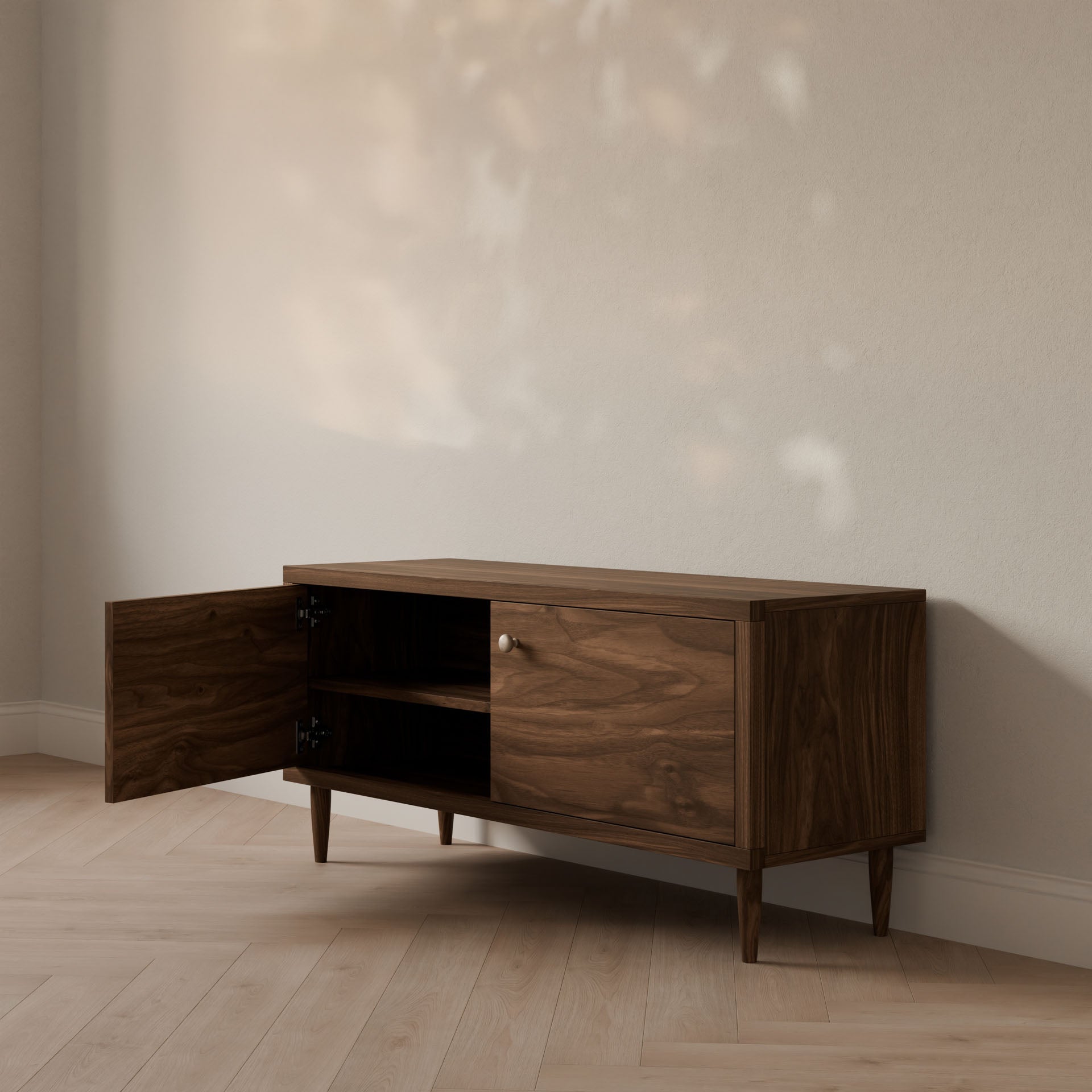The Maine Made Double Door Media Cabinet by Krovel Furniture Co. features tapered legs and sits against a light wall on a wood floor. One door is open, showing shelves—ideal for customizable storage. Soft light falls above, enhancing the scene.