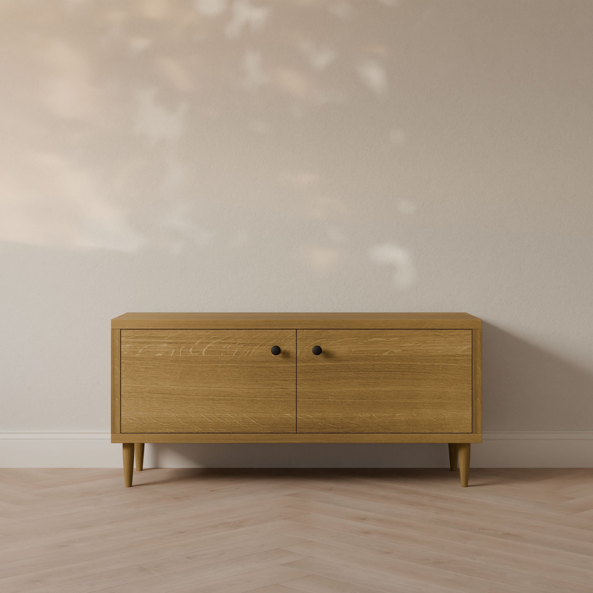 The Maine Made Double Door Media Cabinet by Krovel Furniture Co. features two doors with black knobs, is formaldehyde-free, and stands against a light beige wall on a herringbone-patterned floor—ideal for customizable media setups.