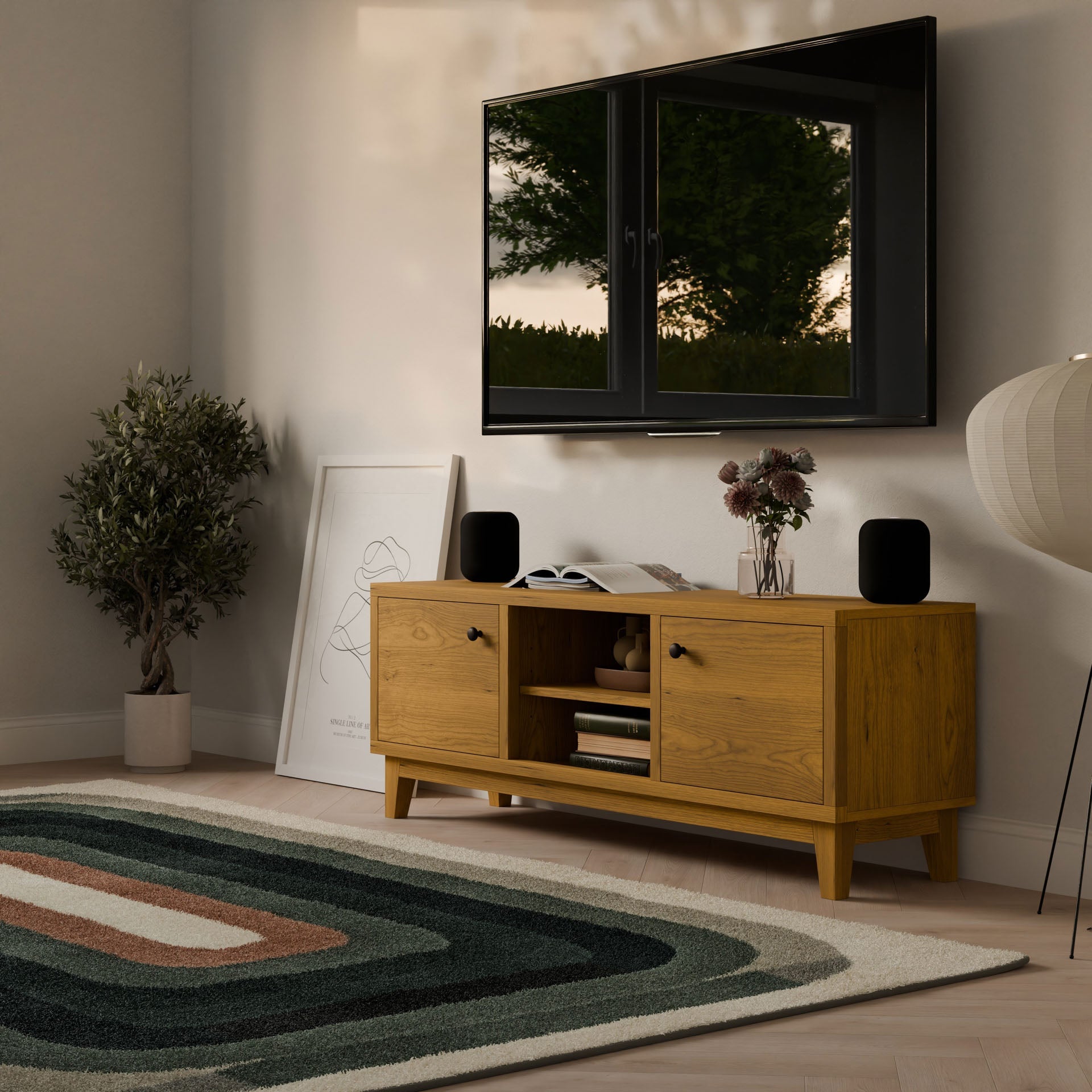 A modern living room features the Maine Made Double Door, Open Shelf Media Cabinet from Krovel Furniture Co., a wall-mounted flat-screen TV, potted plant, abstract art, patterned rug, floor lamp, decor pieces, and a window reflecting trees outside.