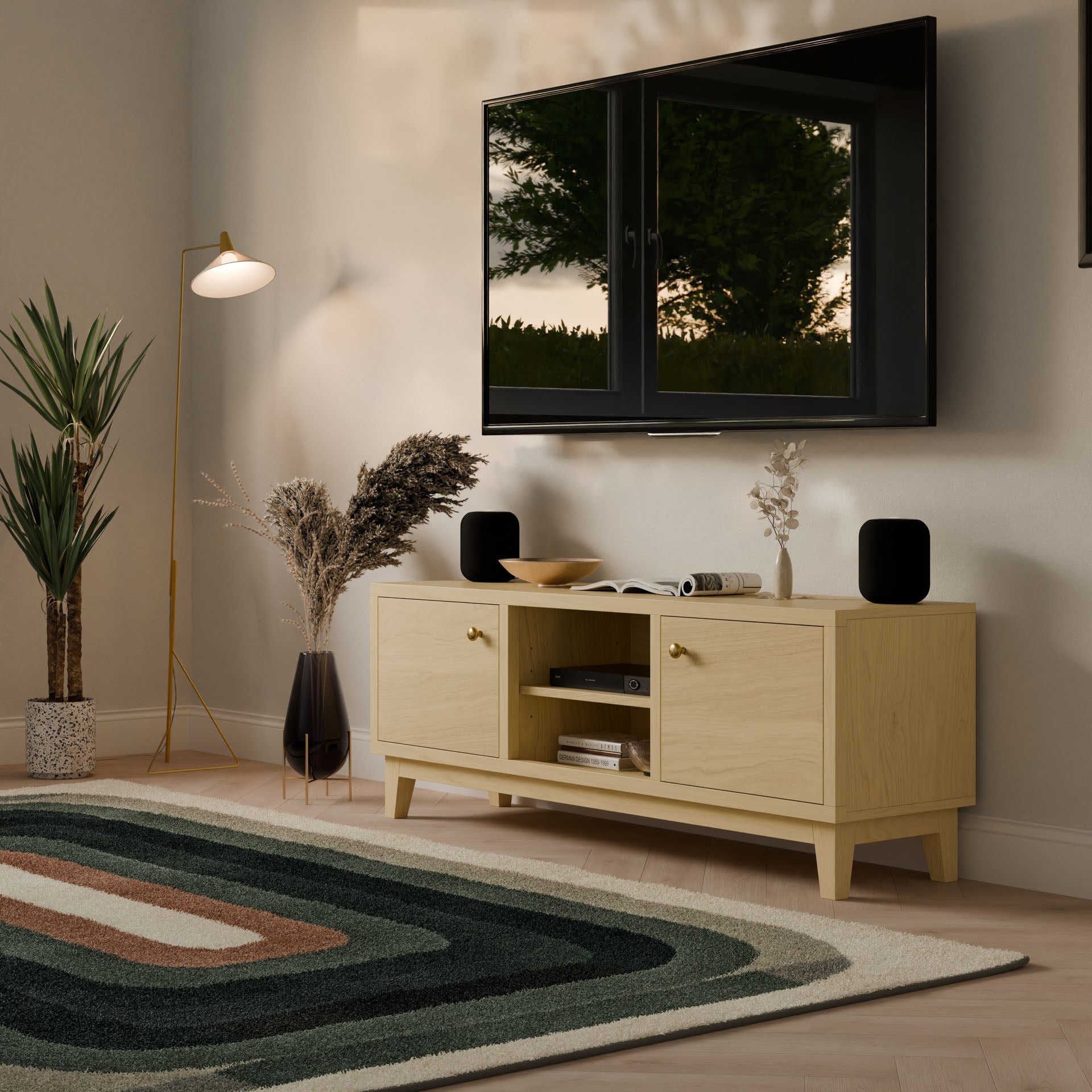 A modern living room with a wall-mounted TV, decorative plants, a floor lamp, an earthy-toned geometric rug, and the Krovel Furniture Co. Maine Made Double Door, Open Shelf Media Cabinet holding books and vases beneath a window reflecting greenery.