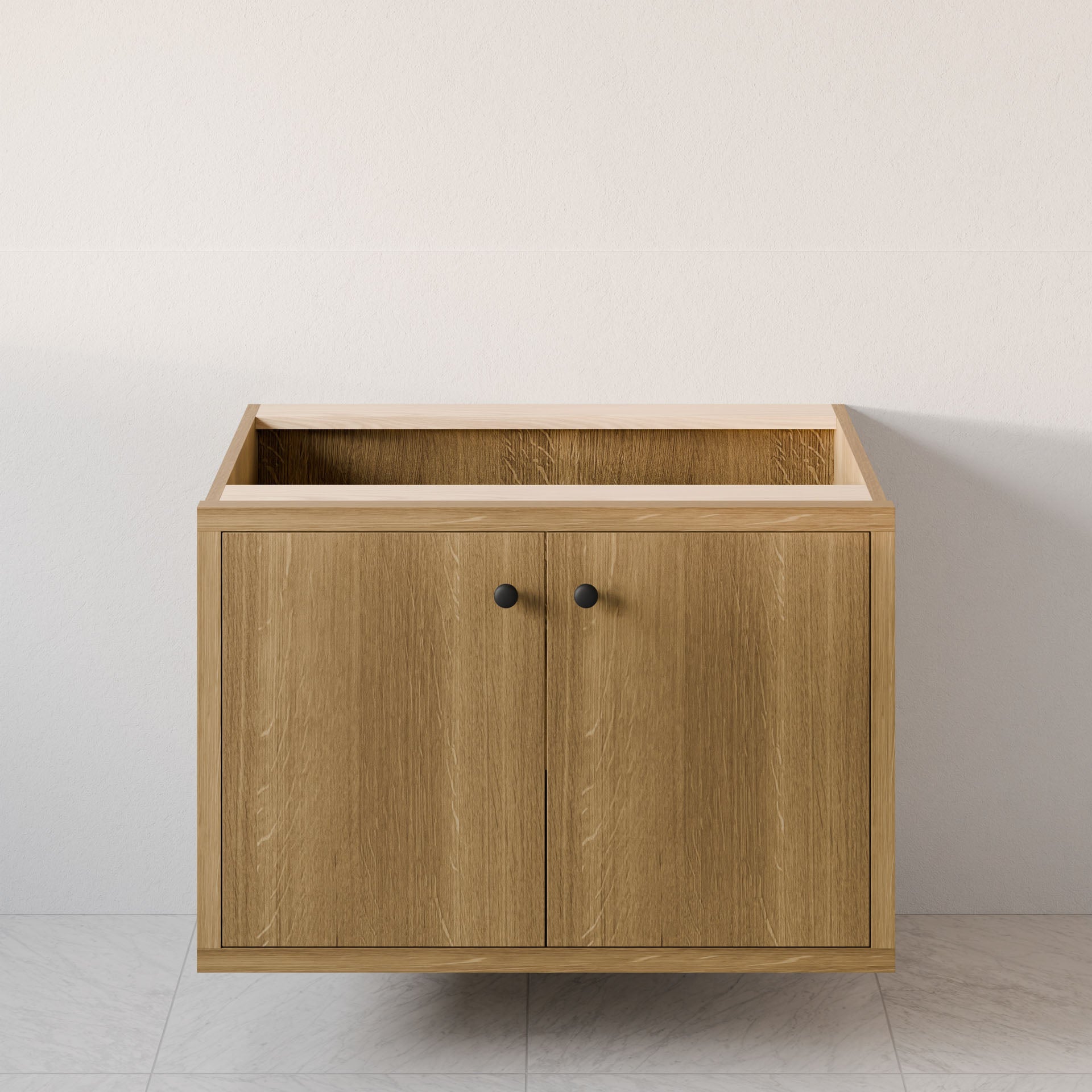 The Maine Made Double Door Vanity by Krovel Furniture Co. is a handmade hardwood cabinet with two doors and black knobs, shown without a countertop or sink, set against a light wall on tiled flooring.