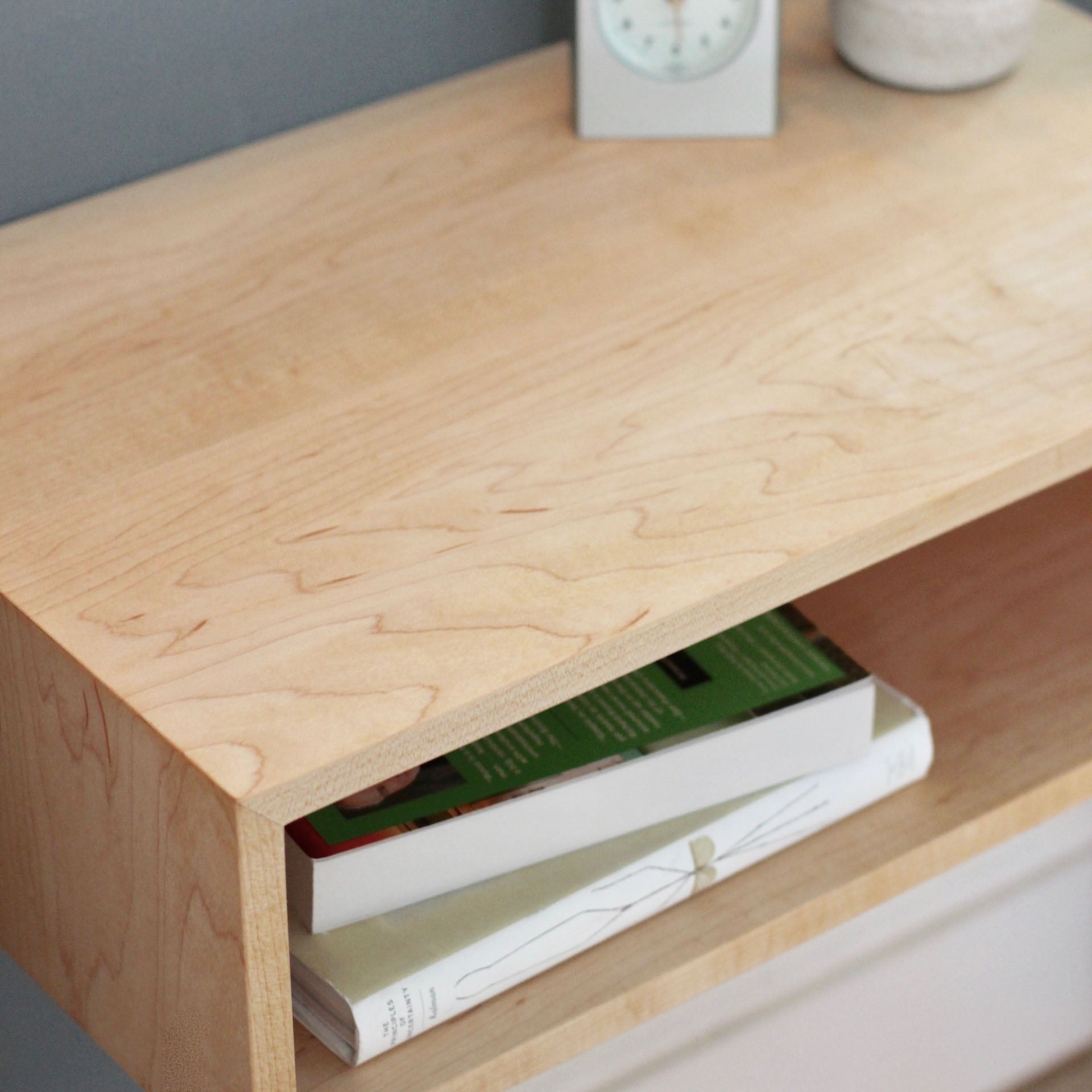 A close-up of the Krovel Hardwood Floating Nightstand Shelf showcases its open shelf with two books, while the surface holds a small round white container and a square clock, all set against a blue-gray wall.