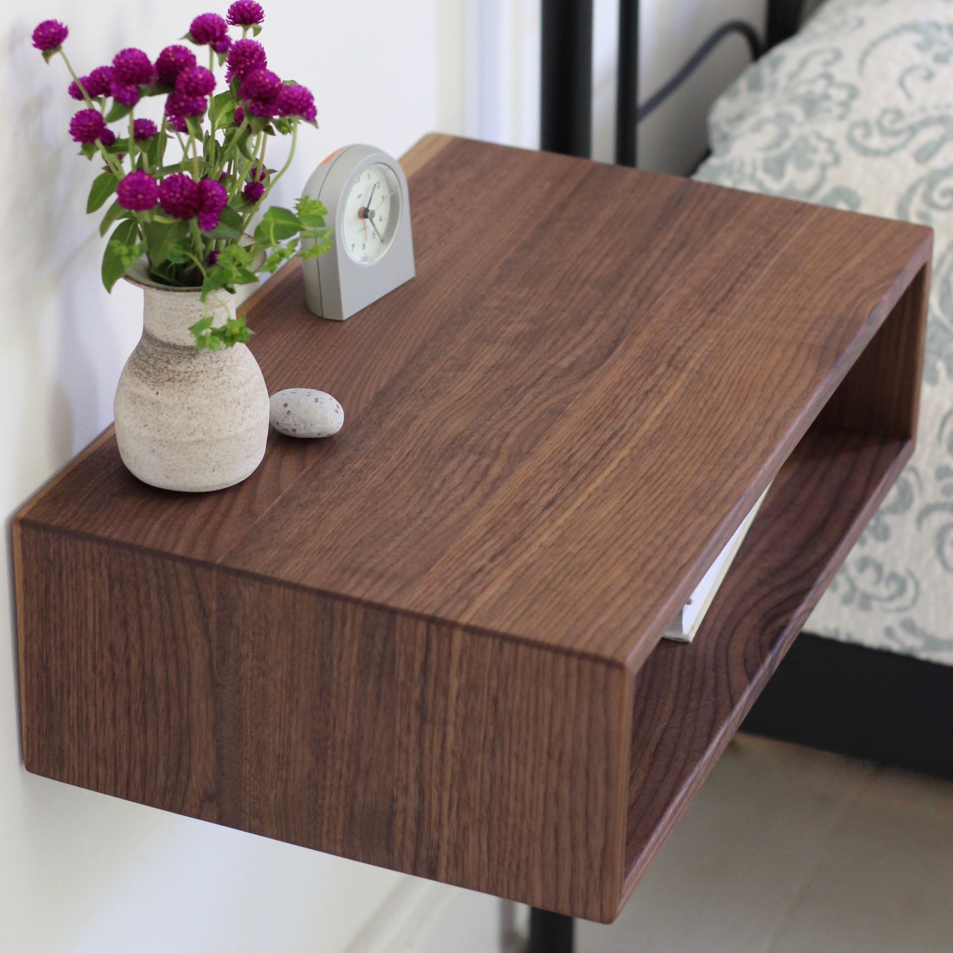 The Krovel Hardwood Floating Nightstand Shelf holds a vase of purple flowers, a small round clock, and a stone beside a bed with a patterned duvet. A book is partly visible on the lower shelf.