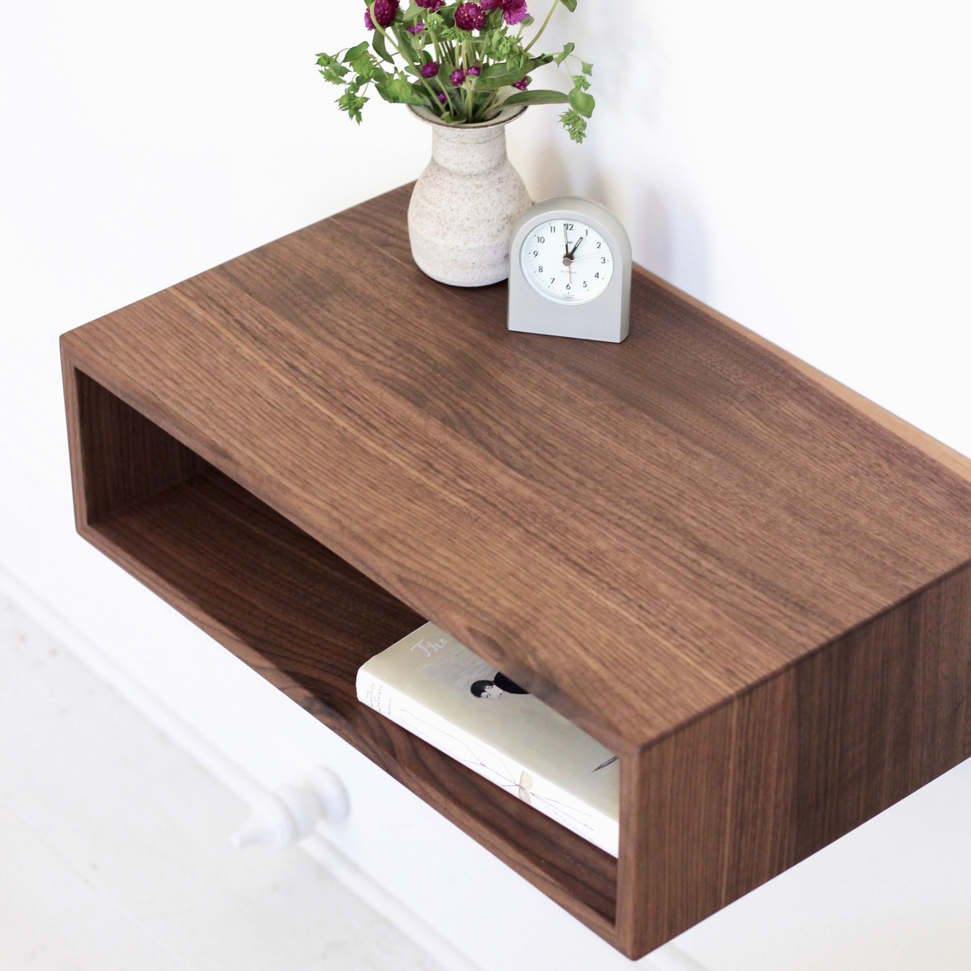 The Krovel Hardwood Floating Nightstand Shelf features an open shelf with a book, topped with a small vase of purple flowers and a gray clock, set against a white wall.