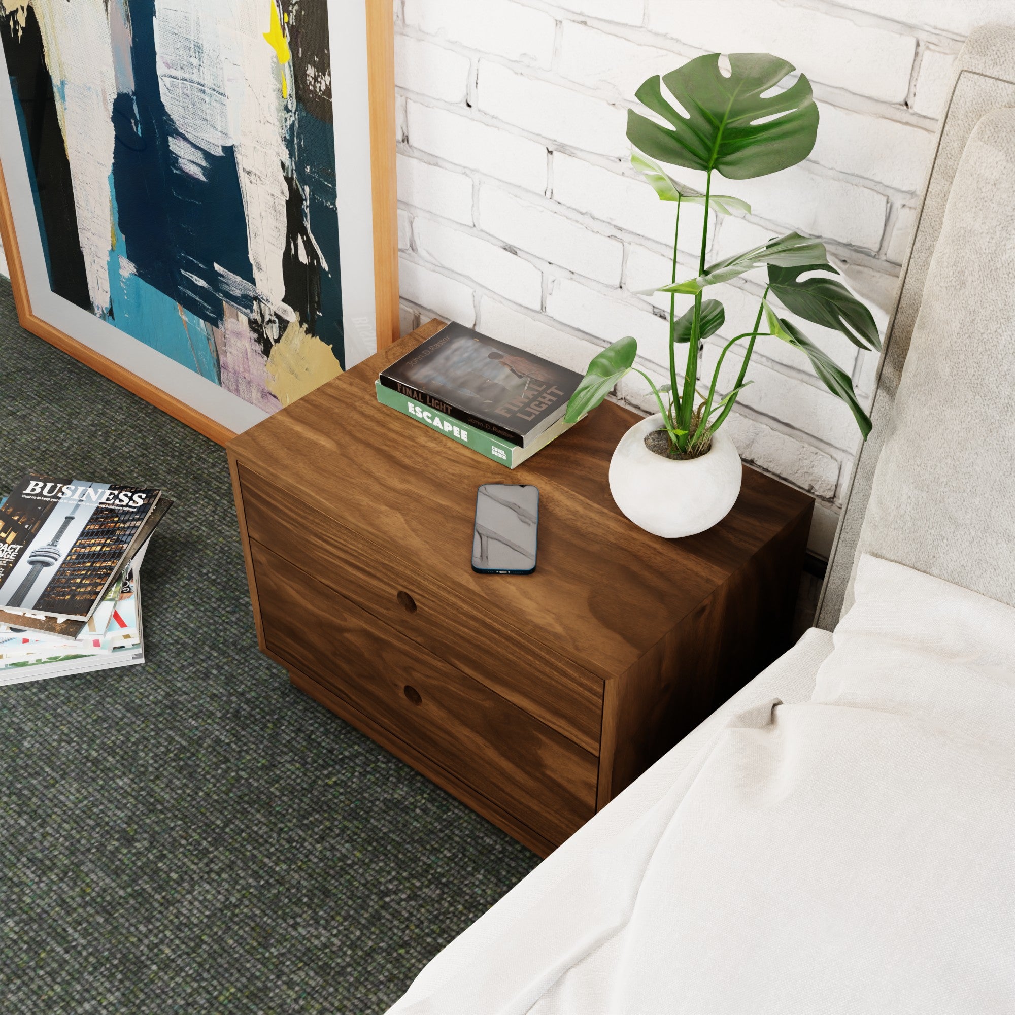 The Krovel Hardwood Freestanding Nightstand with two drawers holds a potted plant, phone, and book. An abstract painting leans against a white brick wall beside the nightstand next to a bed with white bedding. A magazine lies on the floor.