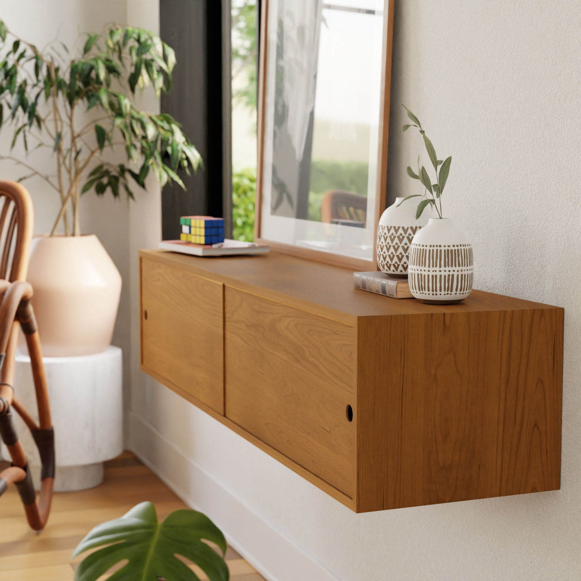 The Krovel Hardwood Oversized Media Console is mounted on a white wall below a framed picture, topped with two patterned vases, greenery, and a Rubik's Cube in a bright room with a nearby plant and chair.