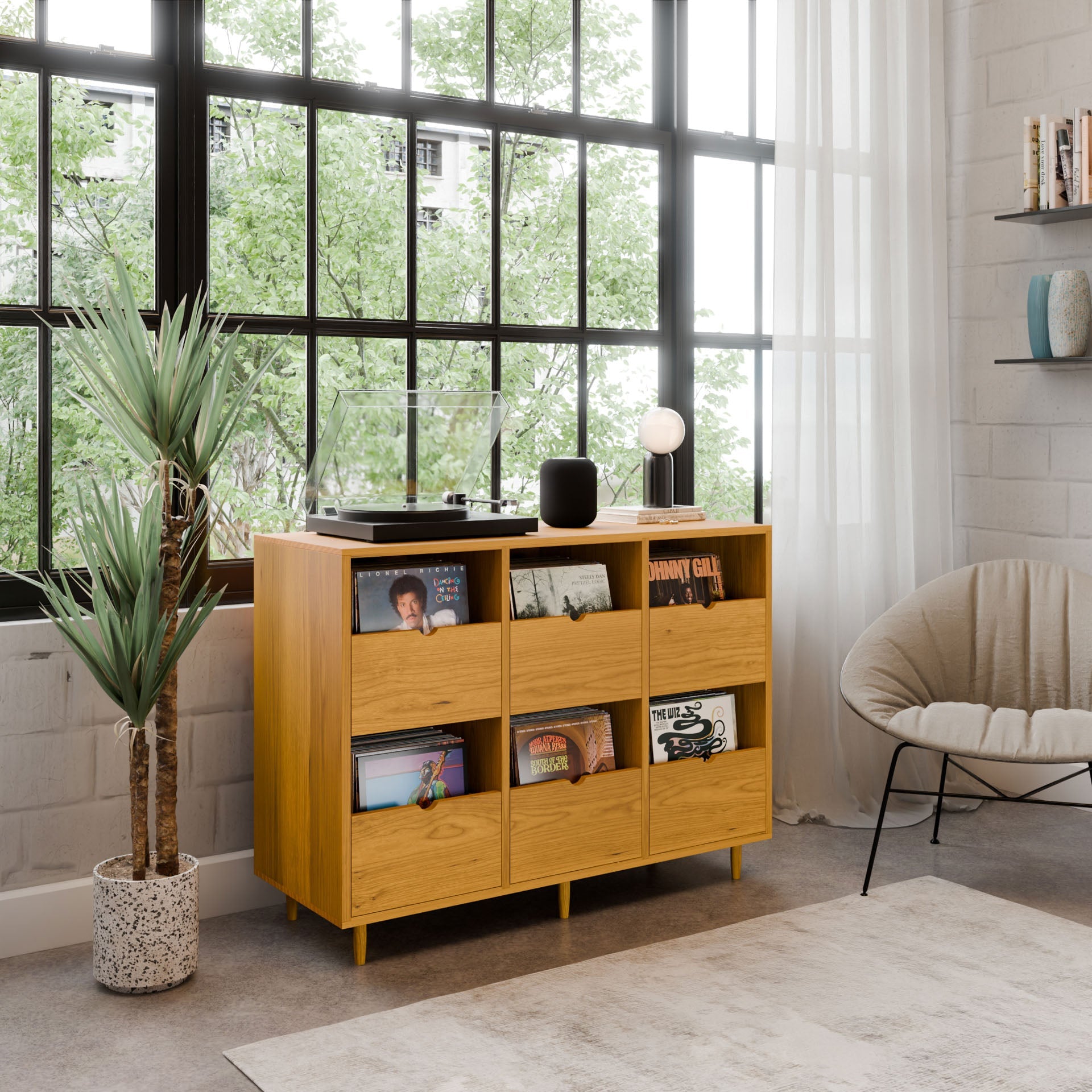 A modern living room with a large window, potted plant, circular chair, and light rug on wooden floors. A Krovel Hardwood Six Drawer Record Credenza holds records and a turntable. Shelves with books hang on the white brick wall.