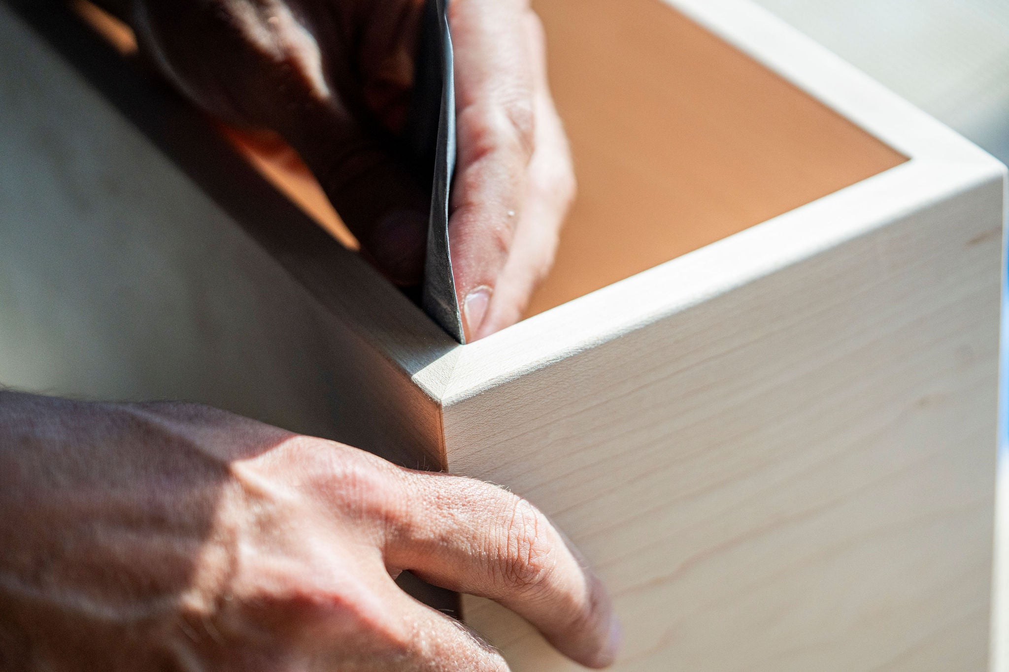 Close-up of hands sanding the corner of a light wooden box, smoothing the edges with sandpaper in natural light.