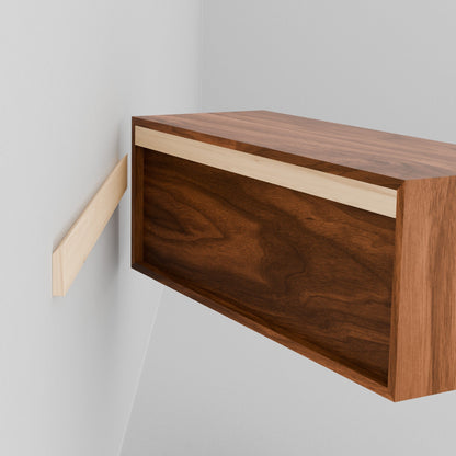 A person aligns a Krovel Hardwood Floating Display Case, featuring a French cleat, with a matching wall-mounted cleat on a white wall, preparing to hang the cabinet.