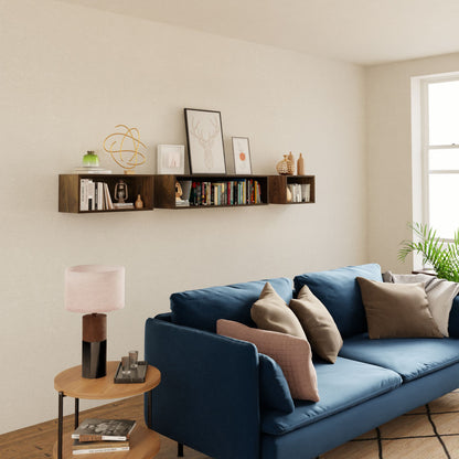 A modern living room features a blue sofa, beige and brown pillows, a round wooden side table with a pink lamp, and the Krovel Floating Bookshelf in Walnut displaying books, art, and decor on a light wall beside a large window.