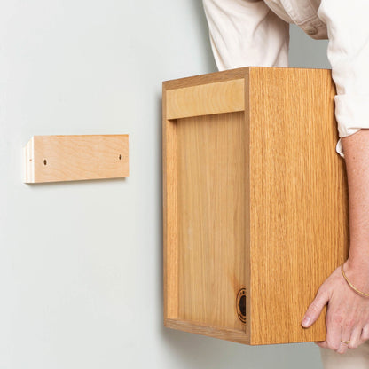 A person installs Krovel Record Storage Shelves in Cherry onto a wall using a rectangular wooden bracket—their hand and arm visible—ideal for vinyl storage or as floating record shelves.