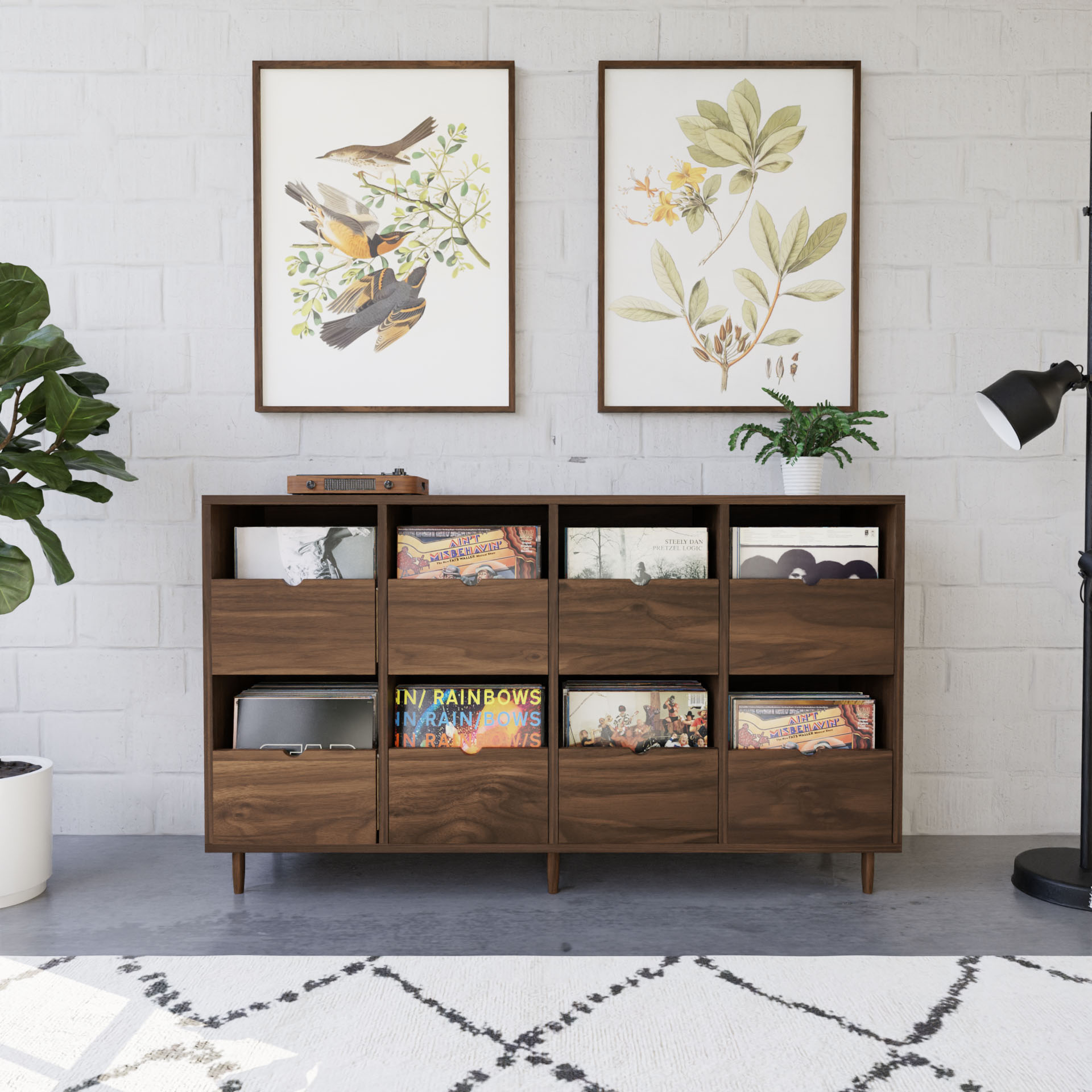 A modern room features a hardwood sideboard, made in the USA, filled with vinyl records under two framed botanical and bird prints on a white brick wall. To the left, a large potted plant, and to the right, a handmade standing black floor lamp graces a patterned rug.