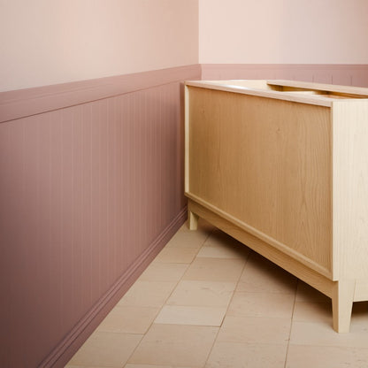 A light natural wood sideboard cabinet in the corner of a room with dusty pink wainscoted walls and beige tiled flooring.