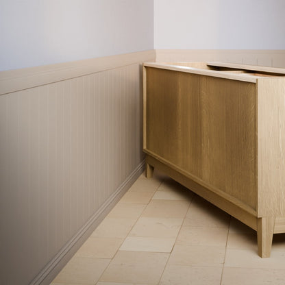 A light oak wood bathroom vanity cabinet with a sink top, shown in a clean, modern bathroom setting with beige tile flooring and wainscoted walls.