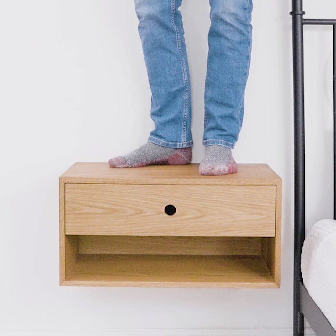A person in jeans and gray socks stands on a hardwood floating shelf with a small drawer. This elegant piece by Krovel Furniture Co. is mounted on a white wall next to a bed frame, embodying the quality of Made in USA craftsmanship.