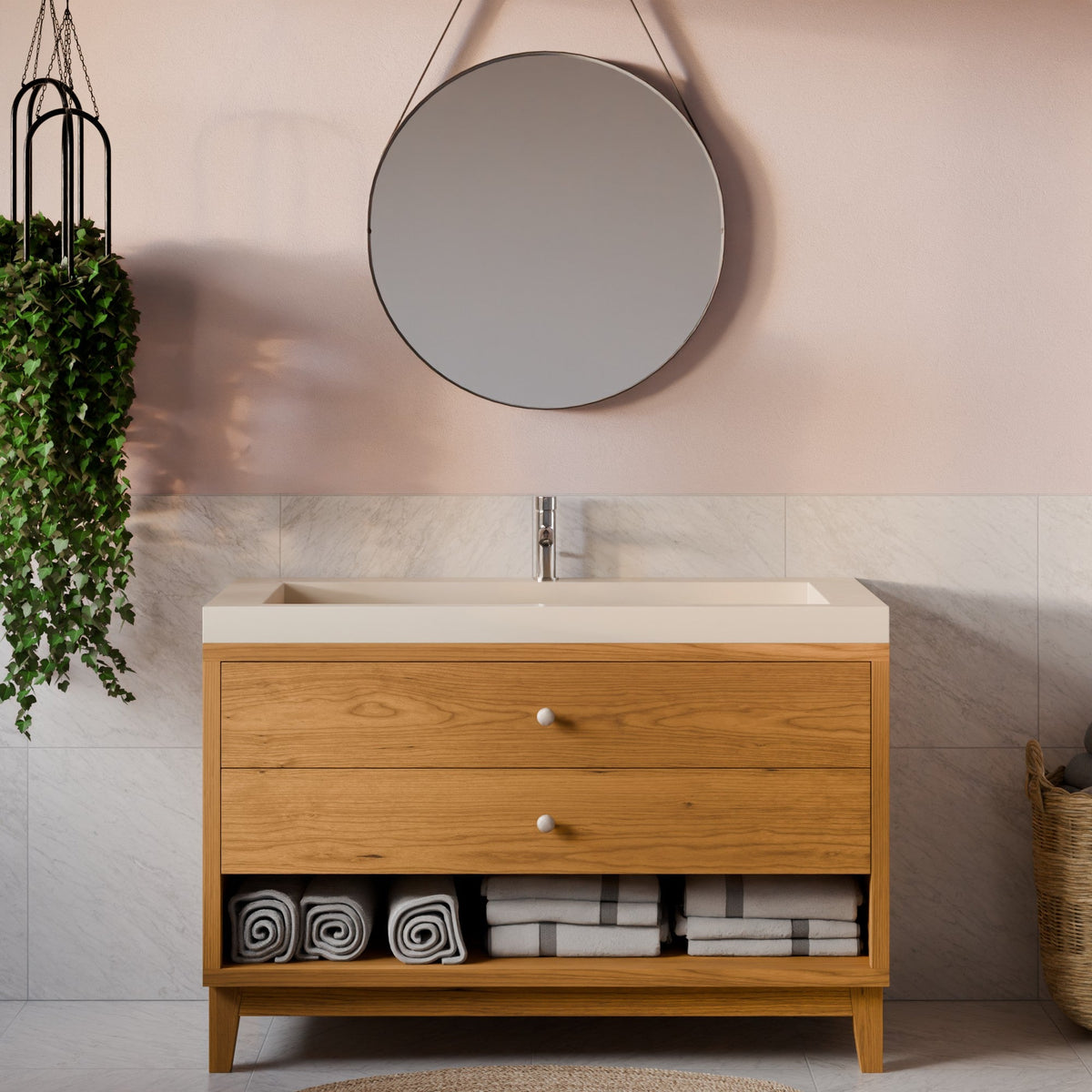 A modern bathroom vanity with a light oak wood cabinet, a cream-colored countertop, and an open lower shelf holding folded towels. A chrome faucet is installed on top, and a round wall mirror hangs on a blush pink wall above.