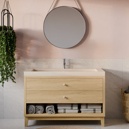 A light oak bathroom vanity with two drawers and an open lower shelf, styled with rolled towels, set against a pink wall with a round mirror.