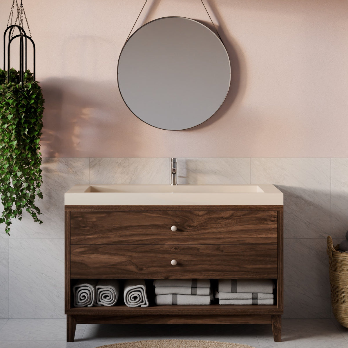 A modern single-sink bathroom vanity with a dark wood cabinet, cream countertop, and open lower shelf, shown in a styled bathroom setting with a round wall mirror, pale pink wall, marble tile, hanging plant, and folded towels.
