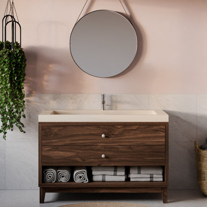 A modern single-sink bathroom vanity with a dark wood cabinet, cream countertop, and open lower shelf, shown in a styled bathroom setting with a round wall mirror, pale pink wall, marble tile, hanging plant, and folded towels.