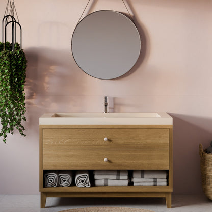 A modern single-sink bathroom vanity with a light oak wood cabinet, cream countertop, and open lower shelf holding folded towels, set against a blush pink wall with a round mirror and hanging plant.