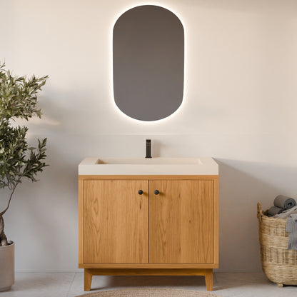 A modern bathroom vanity with a light oak wood cabinet, a white integrated sink countertop, and a matte black faucet. Above it is an oval backlit mirror with a soft white glow.