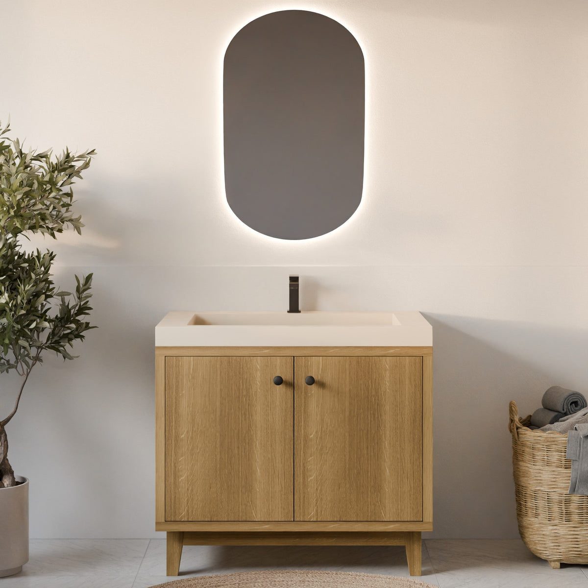 A modern bathroom vanity set featuring a light oak wood cabinet with two doors, a white integrated sink countertop, a matte black faucet, and an oval backlit wall mirror mounted above. The scene is styled with a potted olive tree on the left and a wicker basket on the right against a warm beige wall.