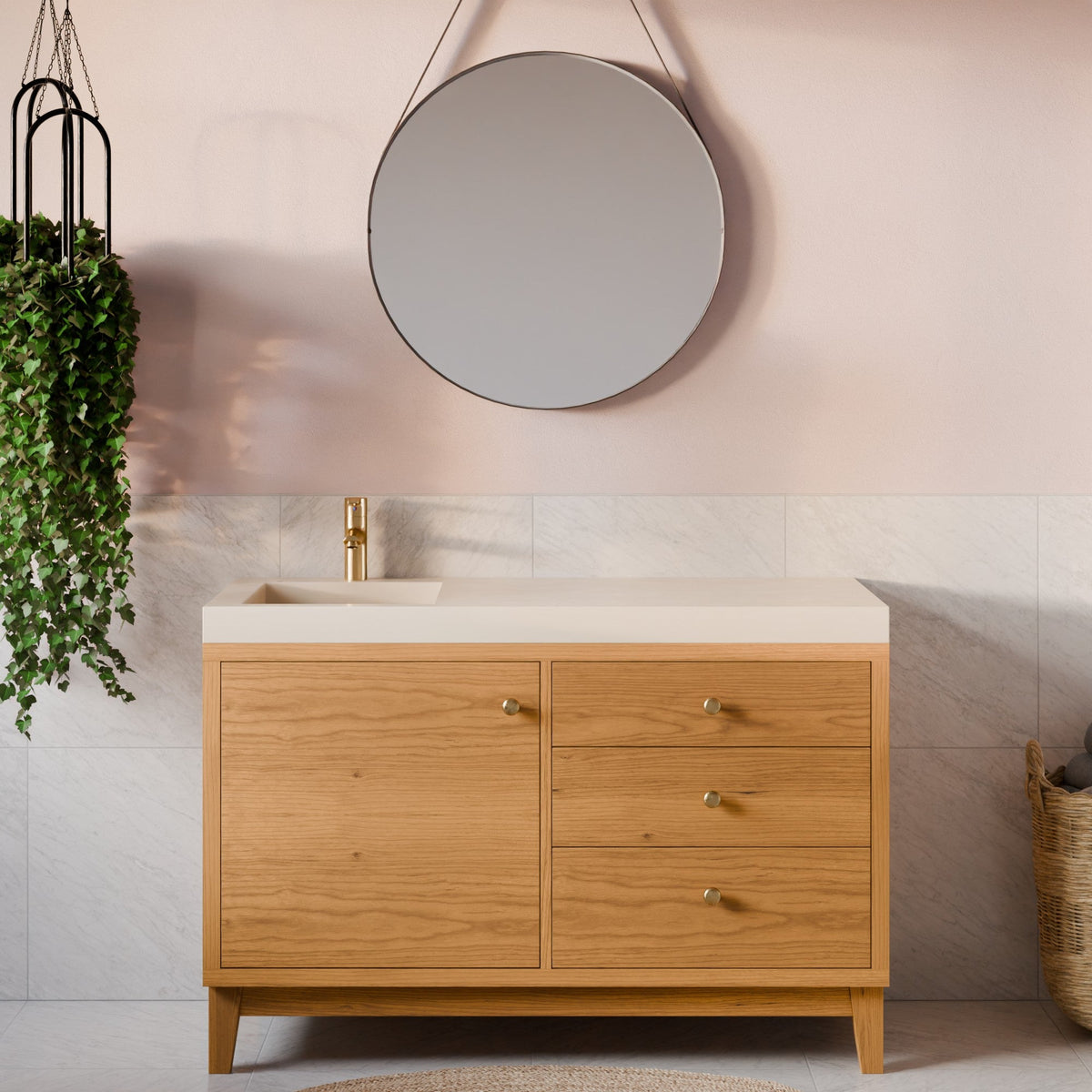 A modern bathroom vanity cabinet with a light oak wood finish, a cream-colored countertop, and a gold faucet, set against a pink wall with a round mirror and a hanging green plant.