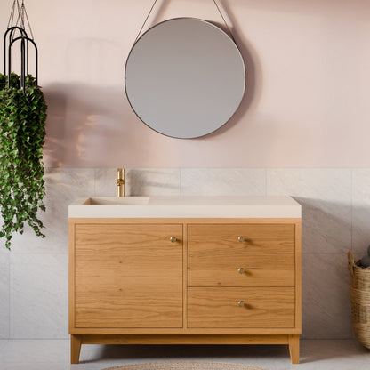 A modern bathroom vanity cabinet with a light oak wood finish, a cream-colored countertop, and a gold faucet, set against a pink wall with a round mirror and a hanging green plant.