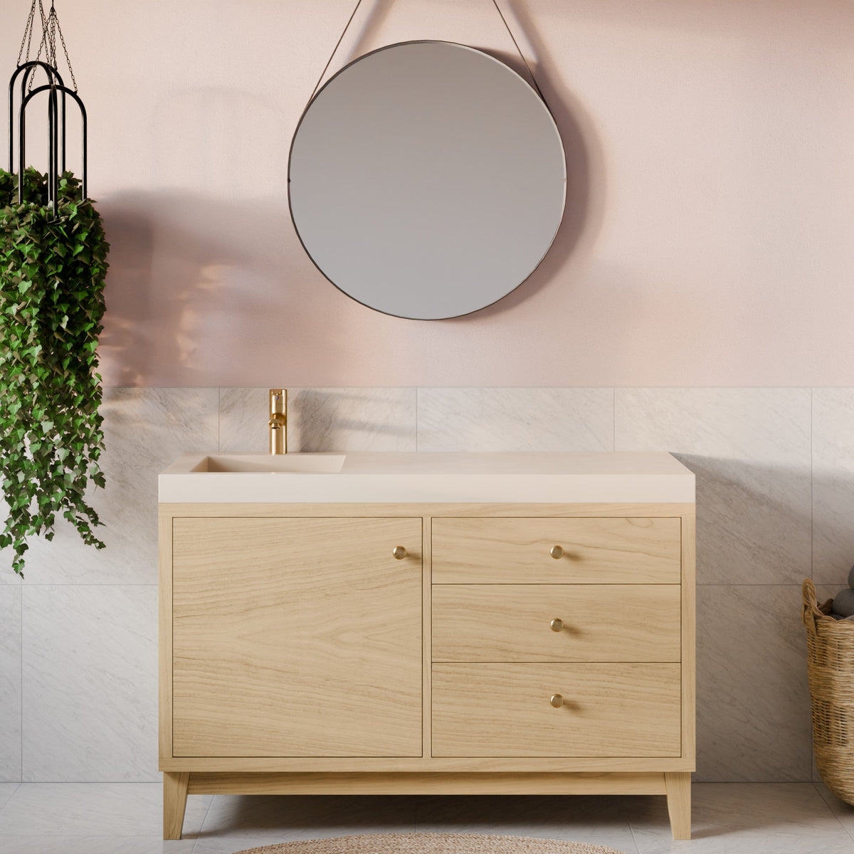 A light oak wood bathroom vanity cabinet with a cream countertop and a gold faucet, set against a pink and white tiled wall with a round mirror and a hanging plant.