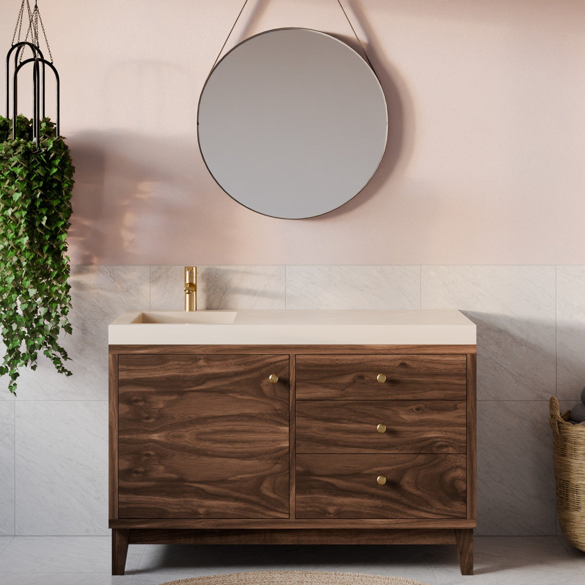 A modern bathroom vanity cabinet with a dark wood grain finish, a cream-colored countertop, and brass hardware, set against a pink wall with a round mirror and a gold faucet.