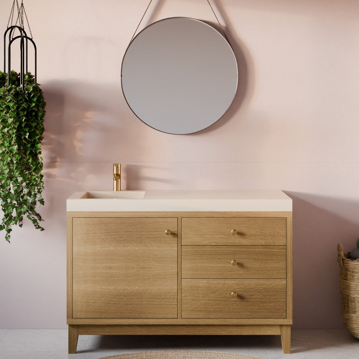 A modern bathroom vanity with a light oak wood cabinet, a white rectangular countertop, and a gold faucet, set against a dusty pink wall with a round mirror and a hanging green plant.