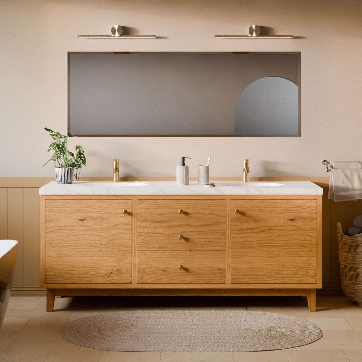 A modern double-sink bathroom vanity with a light oak wood cabinet, white marble countertop, and gold faucets, shown in a styled bathroom setting with a large rectangular mirror, wall sconces, a plant, and a woven rug.