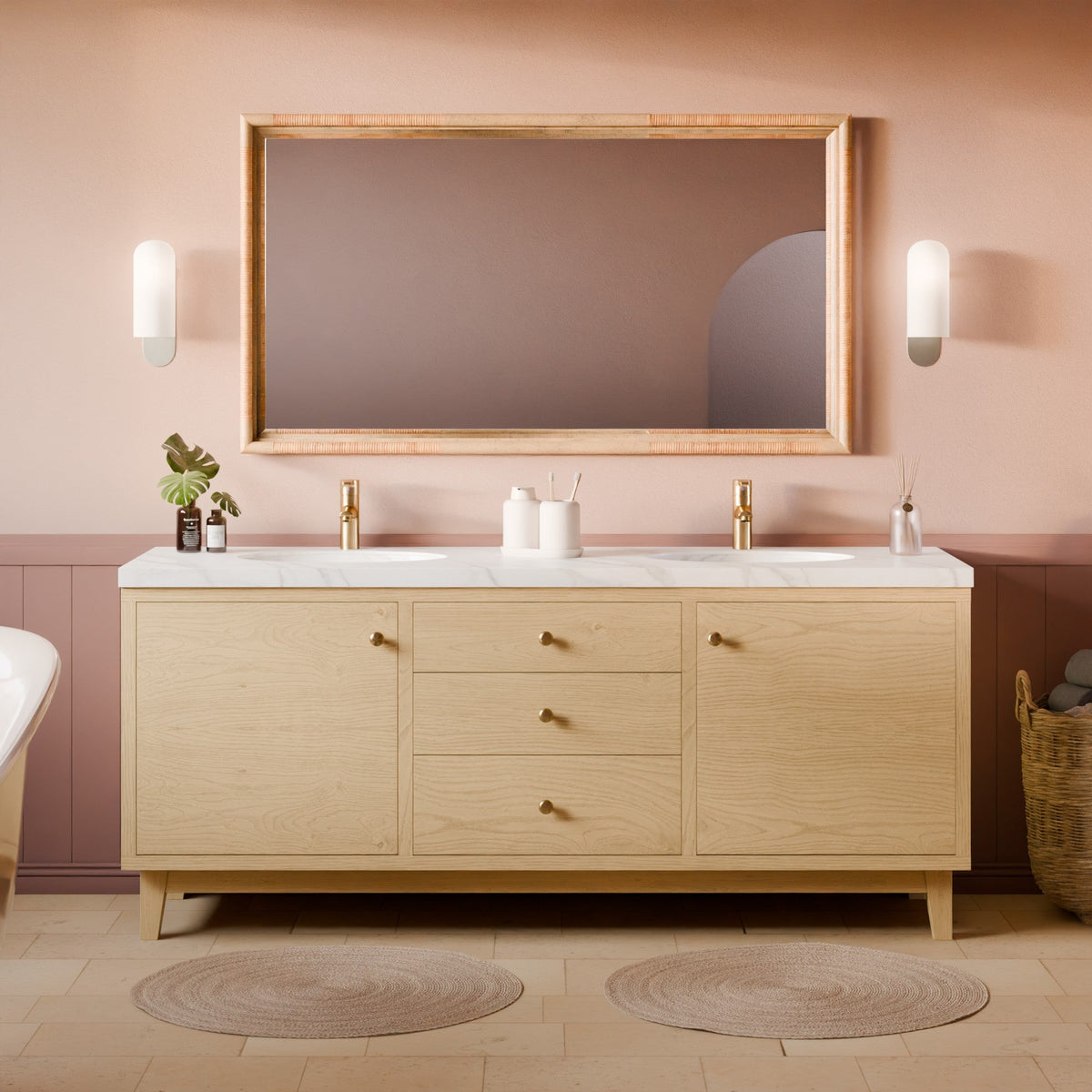 A double-sink bathroom vanity in light natural wood with a white marble countertop, gold faucets, a large rectangular mirror with a wood frame, and a blush pink wall in the background.