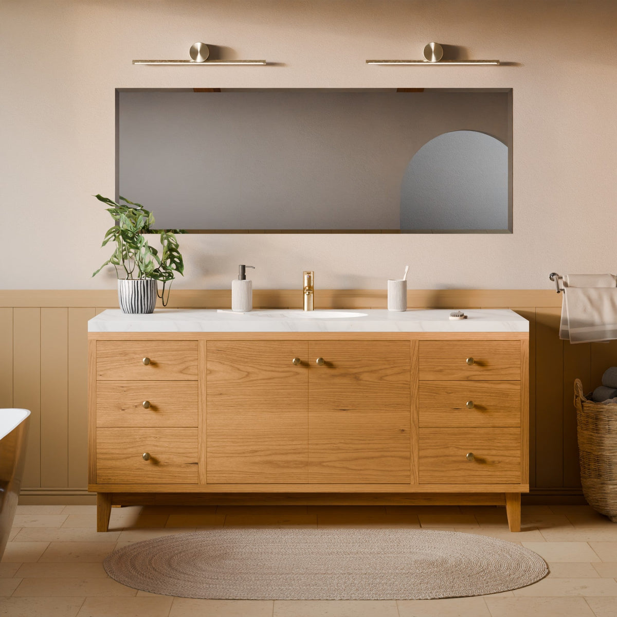 A modern bathroom vanity with a light oak wood cabinet, white marble countertop, and a large rectangular mirror mounted on a beige wall. The vanity is styled with a gold faucet, white soap dispenser, and a potted green plant, set against a warm beige wall with wainscoting.