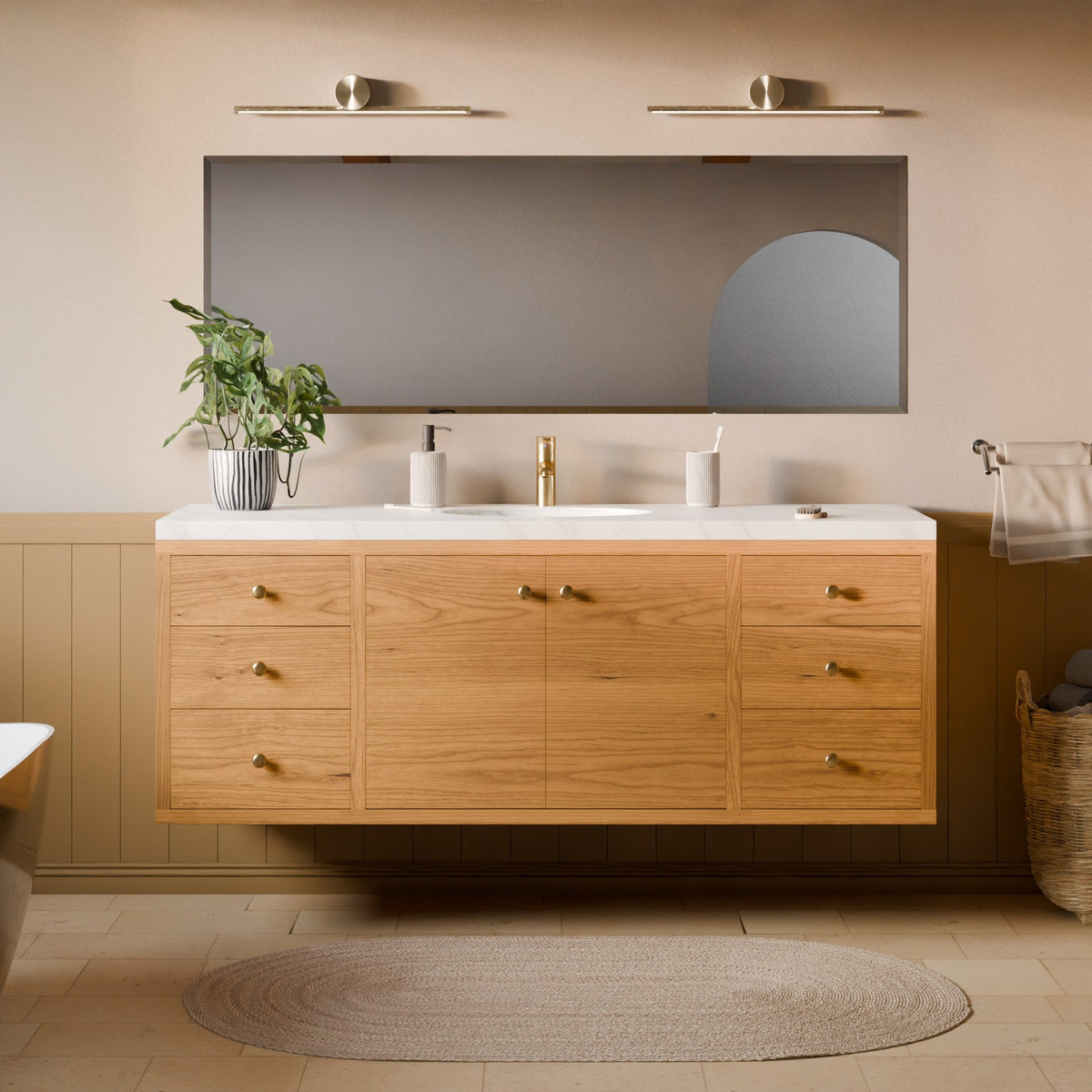 A modern floating bathroom vanity with a light oak wood cabinet, white countertop, and a large rectangular mirror mounted on a beige wall. The vanity is styled in a warm, neutral-toned bathroom with a woven rug, a potted plant, and brass hardware.