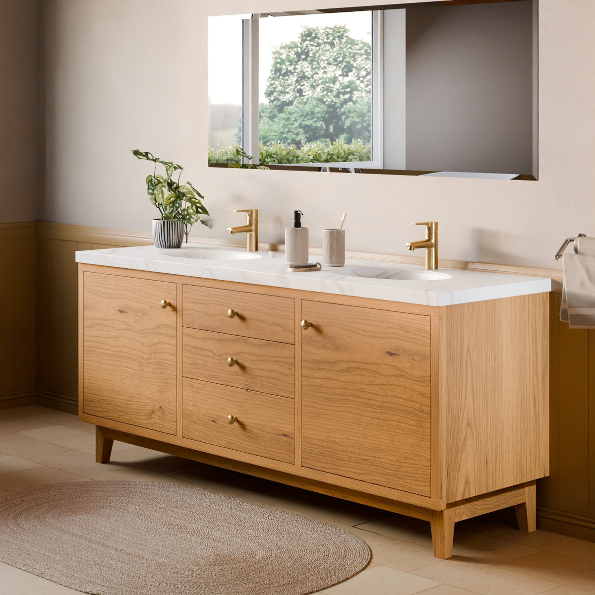 A modern double-sink bathroom vanity with a light oak wood cabinet, white marble countertop, and gold faucets, shown in a styled bathroom setting with a mirror, rug, and window view.