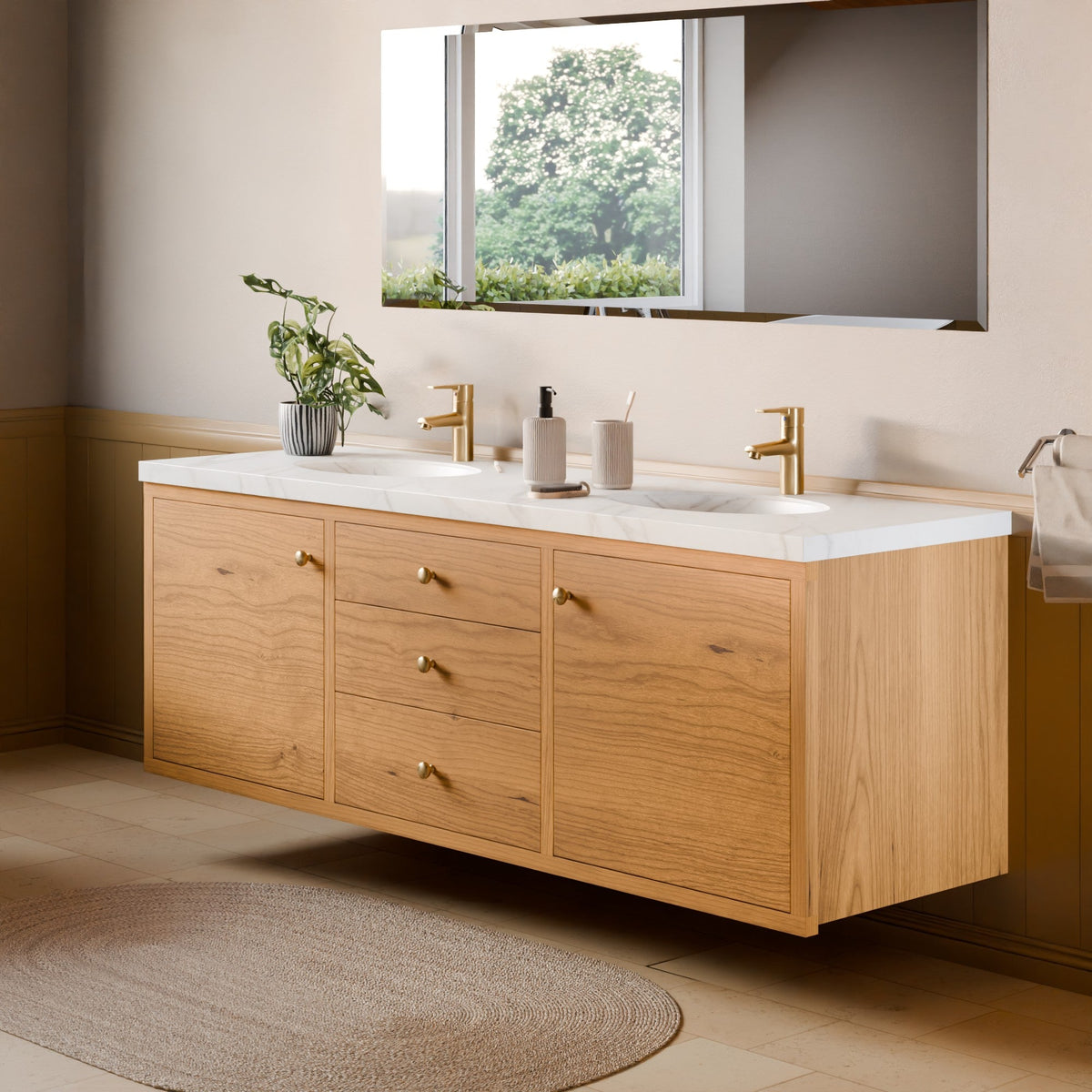 A floating double-sink bathroom vanity made of light natural wood with brass hardware, set against a beige wall with a large mirror and a window above.