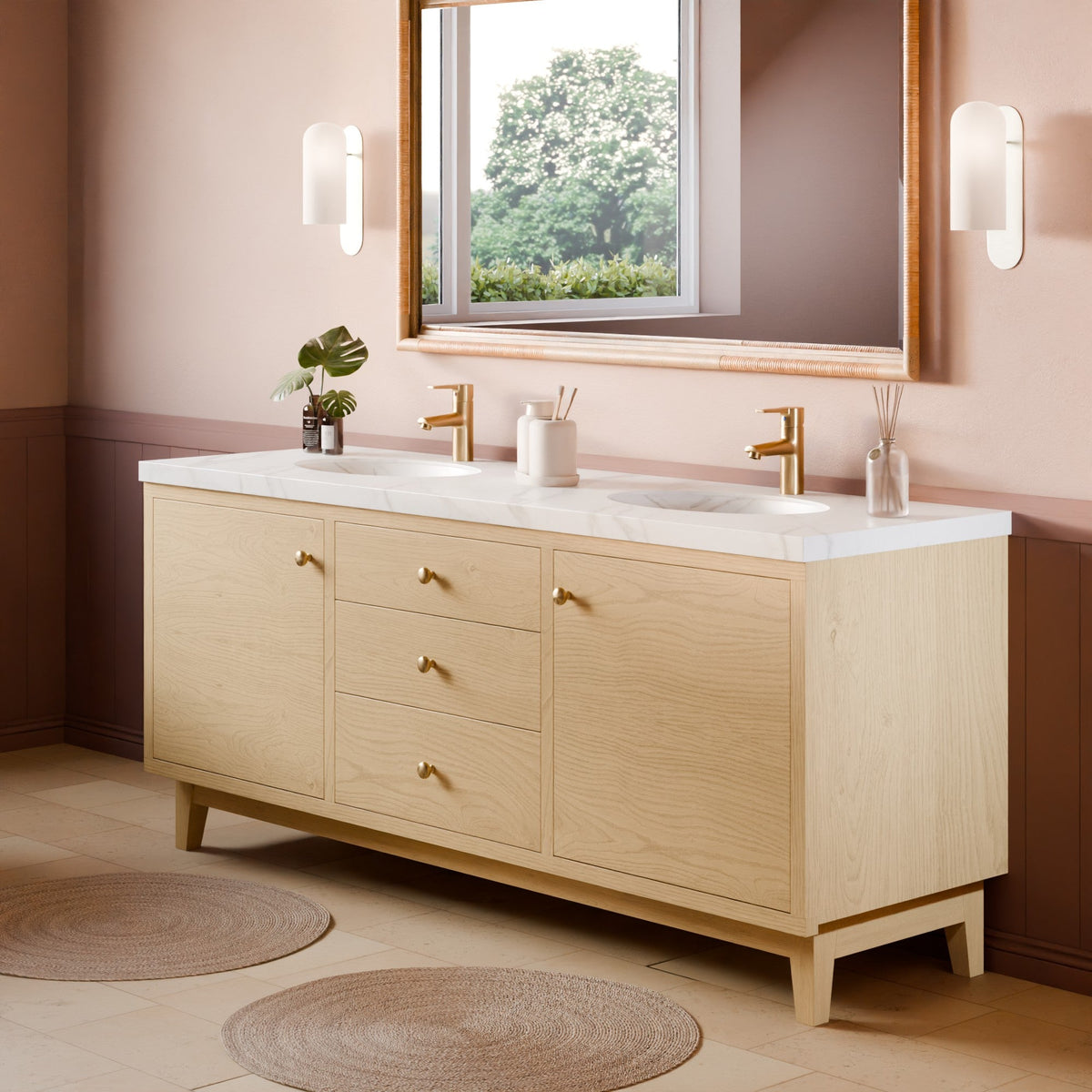 A light natural wood double-sink bathroom vanity with a white marble countertop, gold faucets, and brass-toned knobs, shown in a styled bathroom setting with a mirror, wall sconces, and woven rugs.