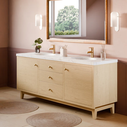 A light natural wood double-sink bathroom vanity with a white marble countertop, gold faucets, and brass-toned knobs, shown in a styled bathroom setting with a mirror, wall sconces, and woven rugs.