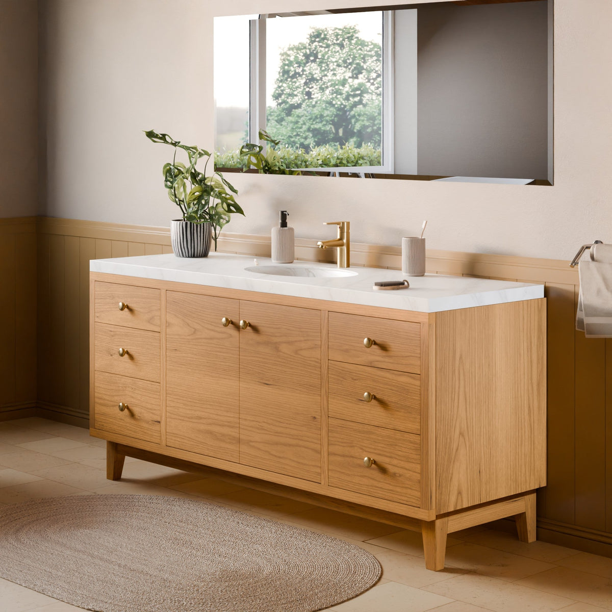 A modern double-sink bathroom vanity with a light oak wood cabinet, white countertop, and gold hardware, shown in a styled bathroom setting with a mirror, plant, and rug.