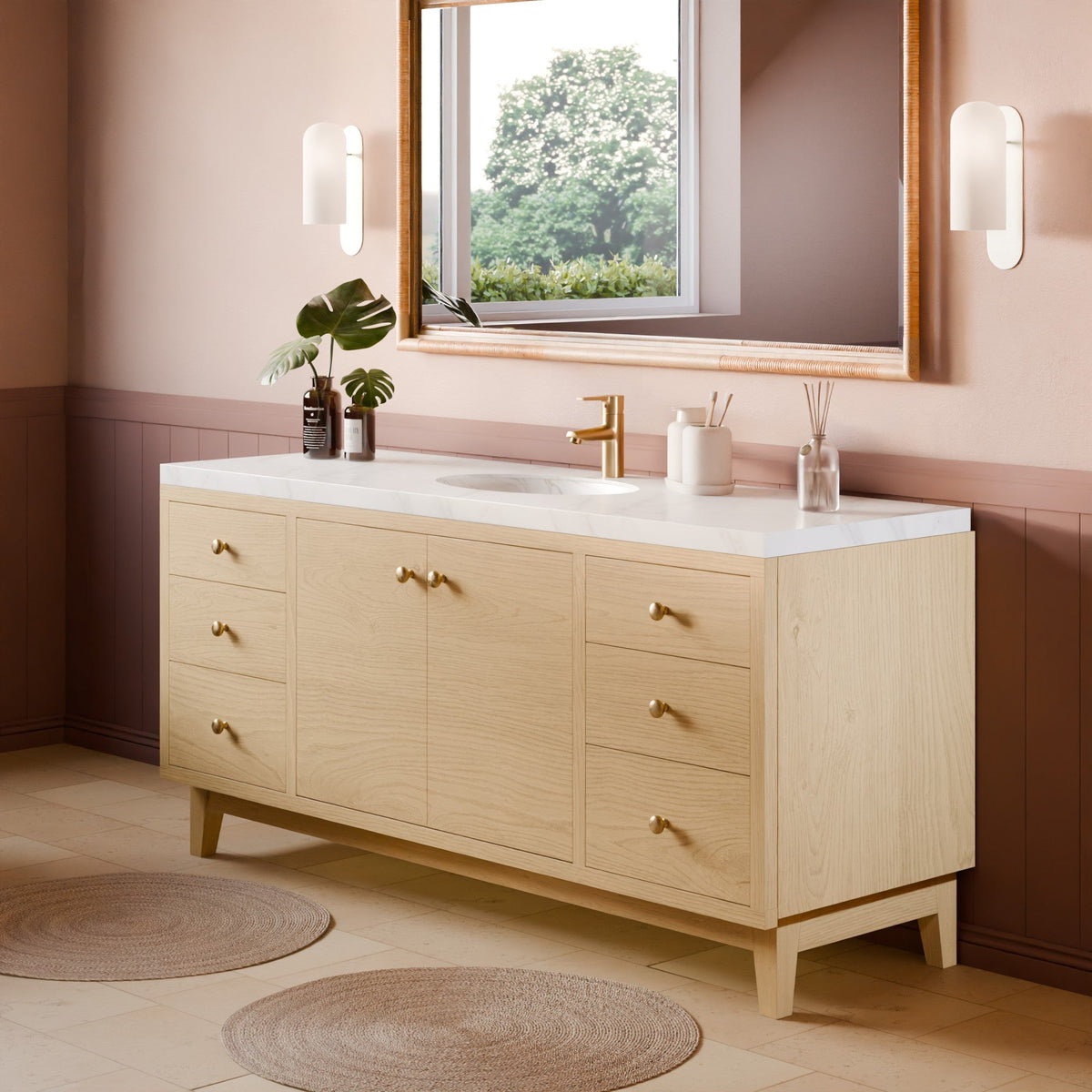 A light natural wood bathroom vanity with a white countertop, integrated sink, and gold faucet, shown in a styled bathroom setting with a mirror, wall sconces, plants, and woven rugs.
