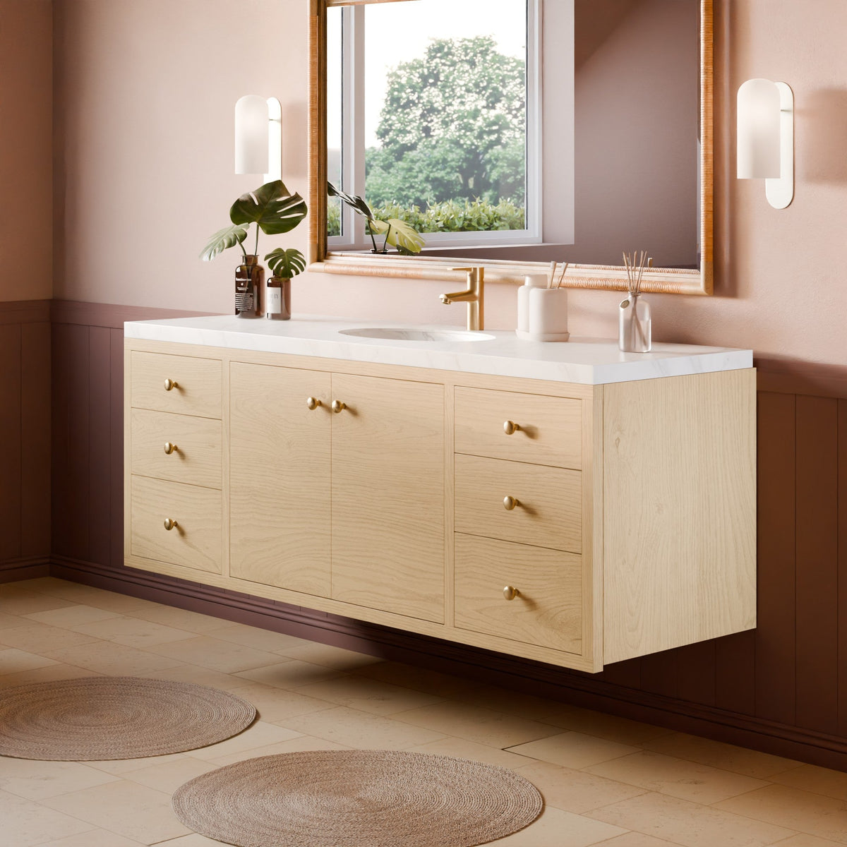 A light natural wood wall-mounted bathroom vanity with a white countertop, gold-tone hardware, and an integrated sink, shown in a styled bathroom setting with a window, mirror, plants, and woven rugs.