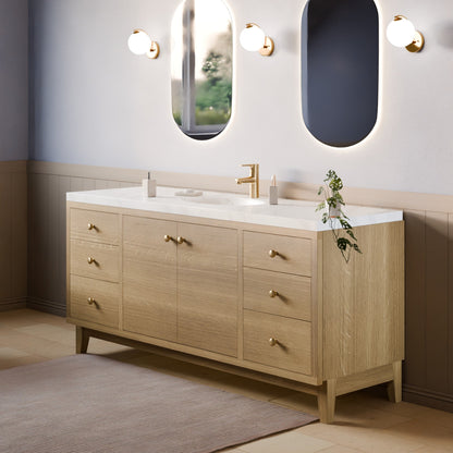 A modern double-sink bathroom vanity in light oak wood with a white countertop, brass hardware, and a gold faucet, shown in a styled bathroom setting with mirrors, wall sconces, and a rug.