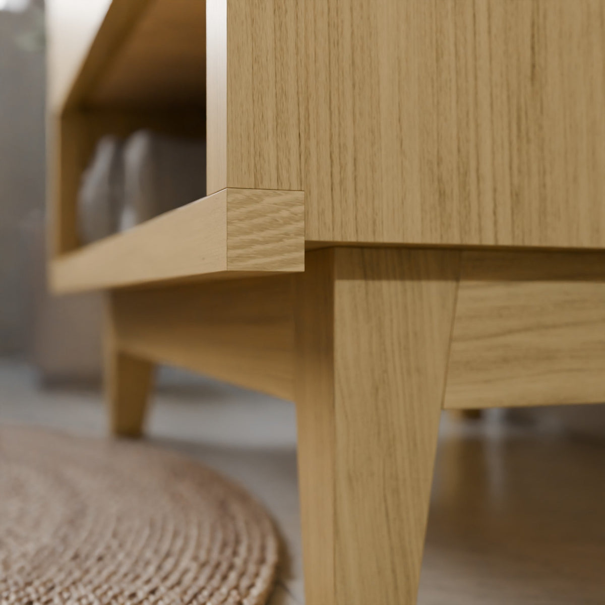 Close-up of a light oak wood furniture piece, showing the corner joint and leg detail against a softly blurred indoor background.