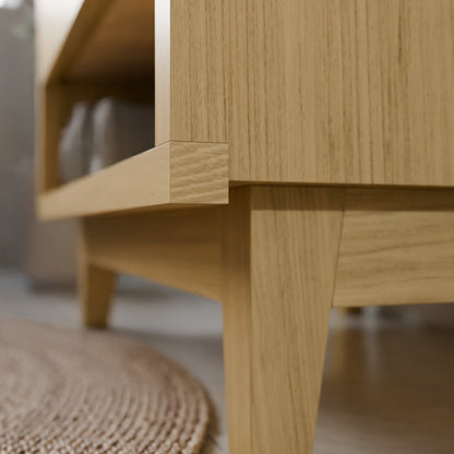 Close-up of a light oak wood furniture piece, showing the corner joint and leg detail against a softly blurred indoor background.