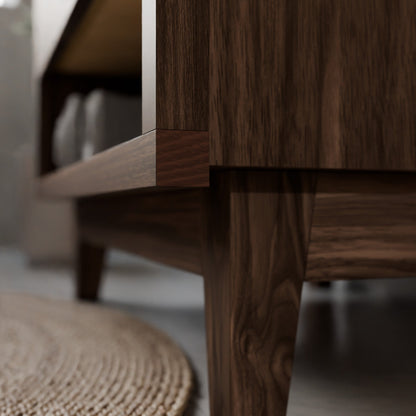Close-up of a dark brown wooden furniture piece, showing the grain of the wood on the leg and side panel, with a blurred woven rug on the floor.