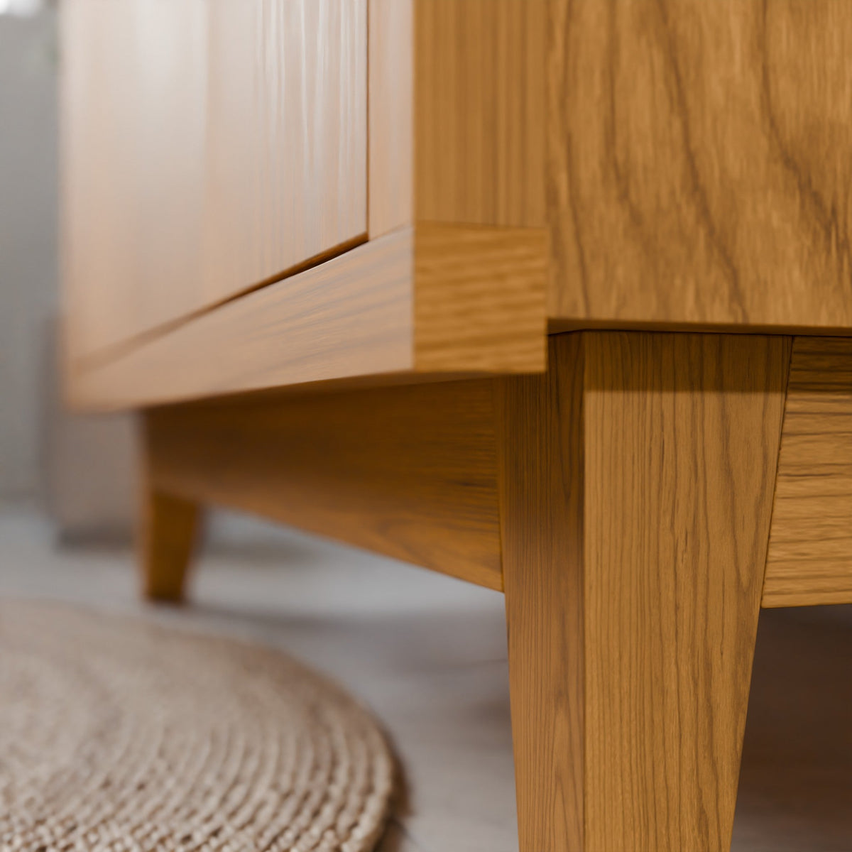 Close-up of a light oak wood furniture piece, showing the corner joint, leg, and cabinet door with a neutral background and a woven rug in the foreground.